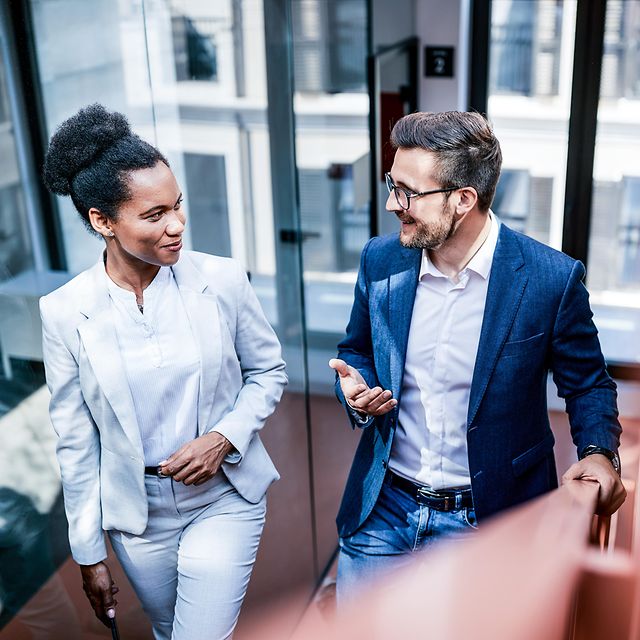 A woman and a man in business attire chat casually as they climb a staircase.