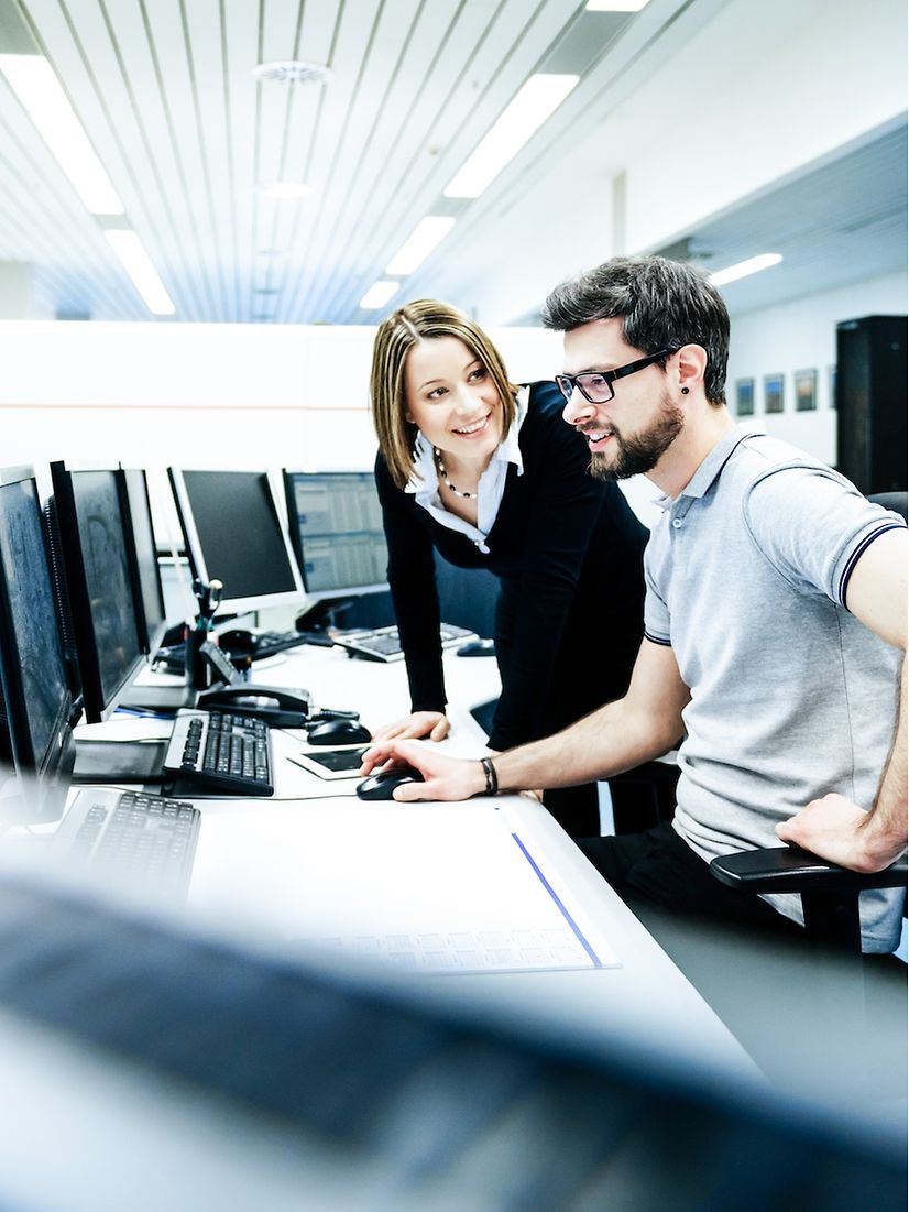 Two people, a woman and a man, are talking to each other at a desk with lots of monitors.