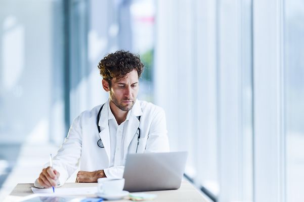 Doctor sits in front of his laptop in his practice and uses the components of Telekom's specialist applications for practices.
