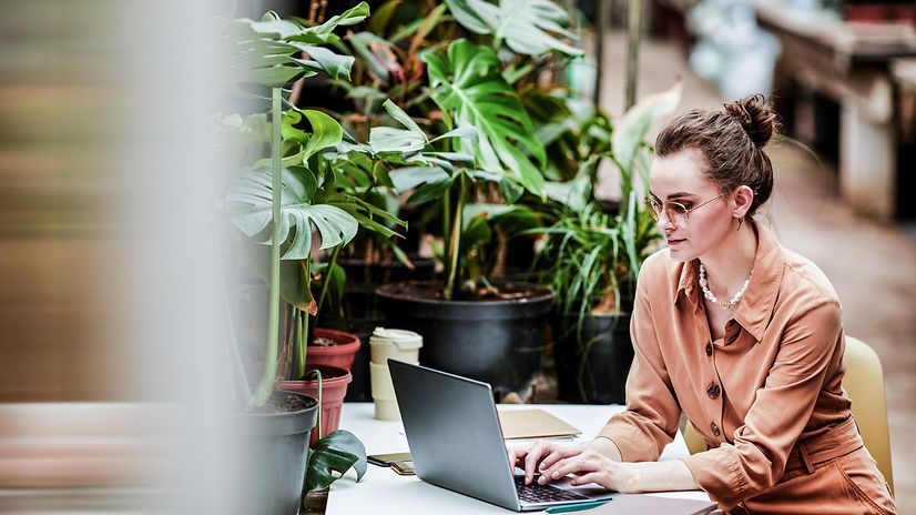A woman sits in front of plants and looks at her laptop.
