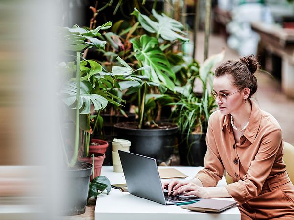 A woman sits in front of plants and looks at her laptop.