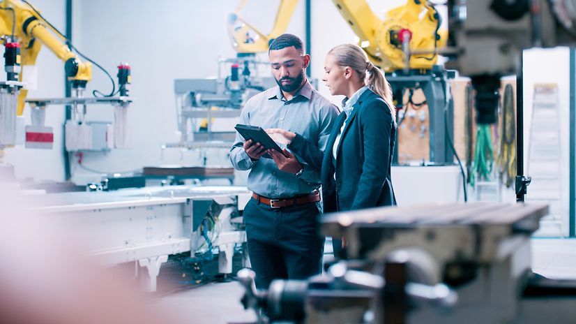Two engineers are standing in a modern production facility, discussing something on a tablet.