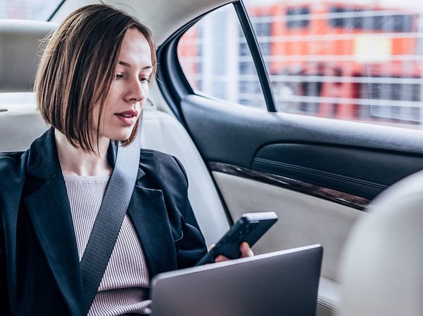 A woman sits in her car with her laptop and smartphone, looking at the screen.