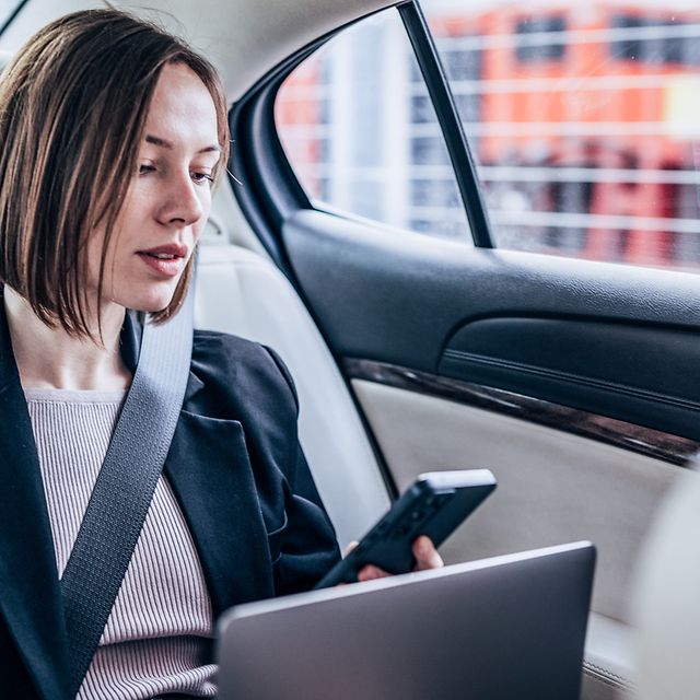 A woman sits in her car with her laptop and smartphone, looking at the screen.