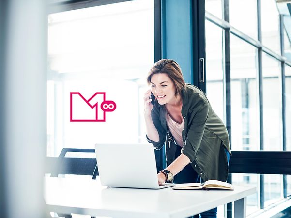 Woman talking on the phone and working on her laptop in a modern office, MagentaEINS UNLIMITED logo in the background.