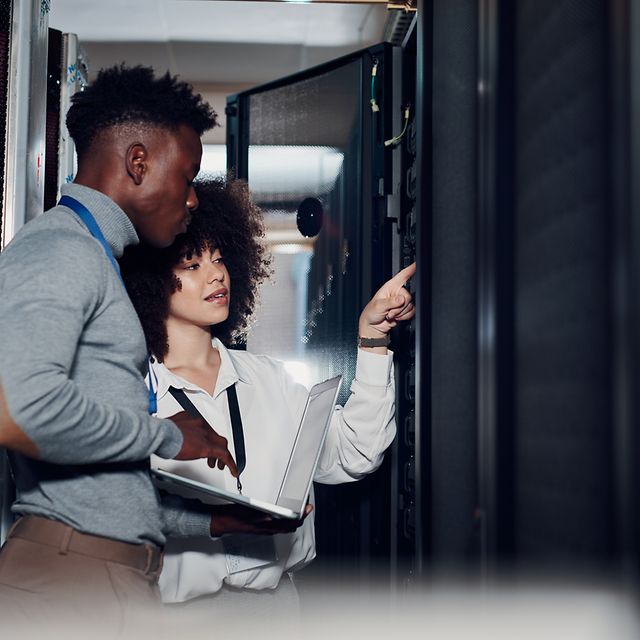 Two network engineers are standing in front of a server cabinet discussing Cisco routing.
