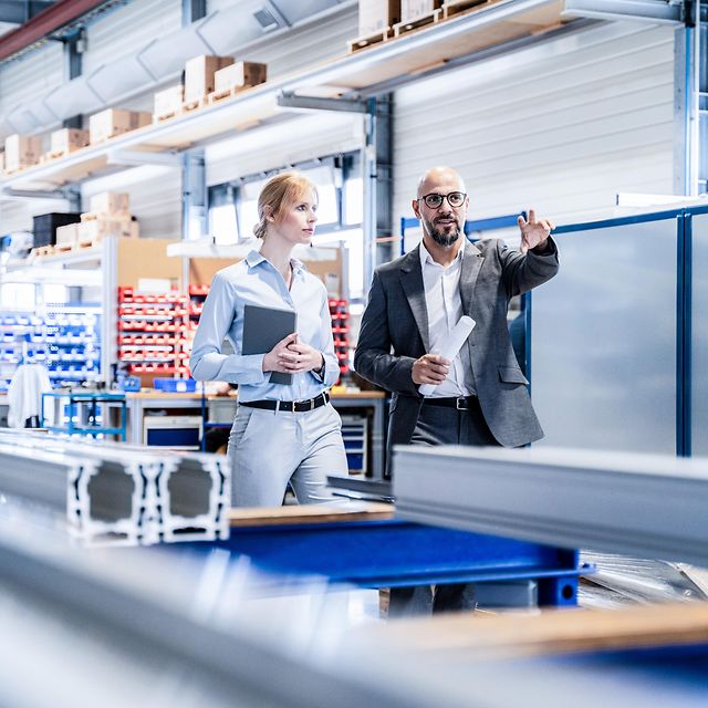 A woman and a man in business attire are discussing matters in a warehouse.
