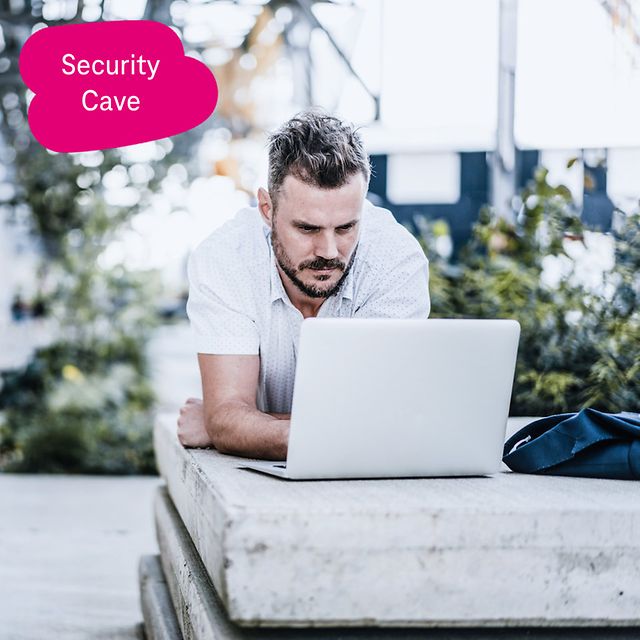 Person arbeitet im Freien liegend mit einem Laptop auf einer Betonkante, im Hintergrund grüne Pflanzen.
