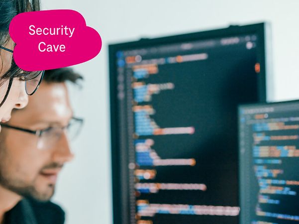 Zwei Personen analysieren Quellcode auf großen Monitoren, eine zeigt mit dem Finger auf den Bildschirm.