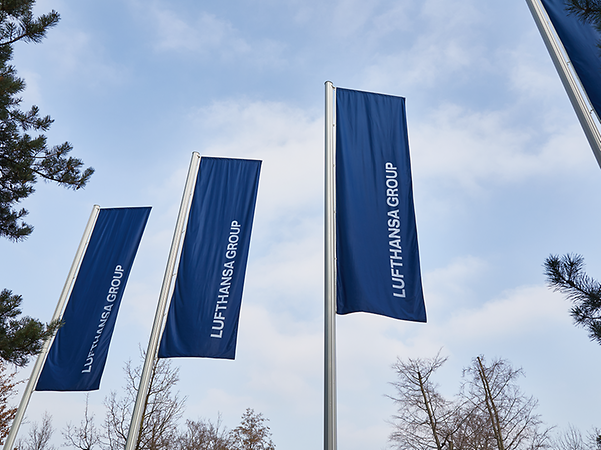 drei blaue Fahnen mit weißem Schriftzug Lufthansa wehen im Wind an Fahnenmasten vor blauem Himmel