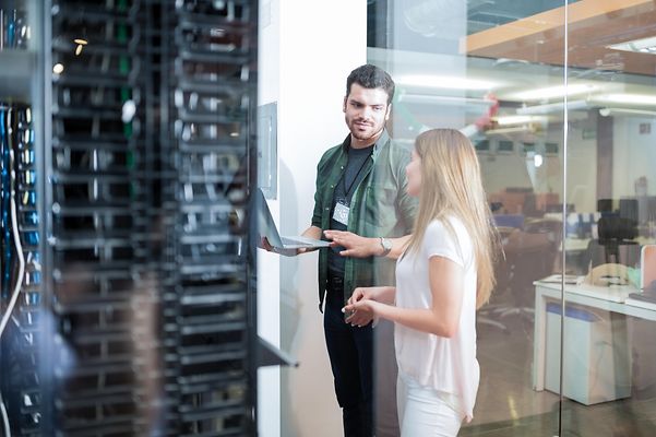 Mann mit Laptop und Frau stehen vor Rechenzentrum mit Cloud-Servern.