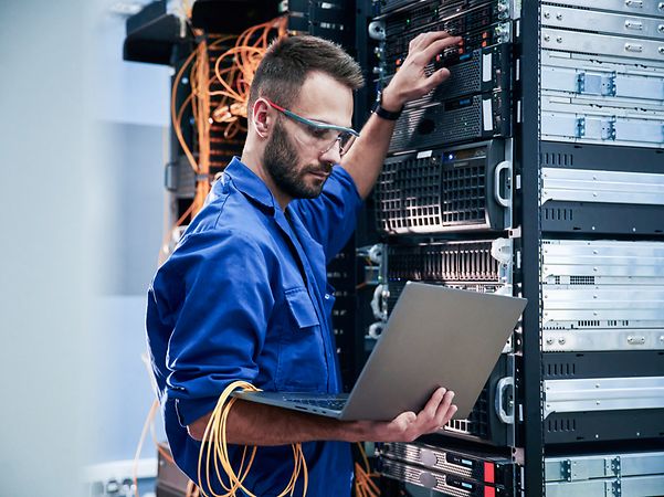 Junger Mann mit Laptop arbeitet mit Internetgeräten und Kabeln im Serverraum