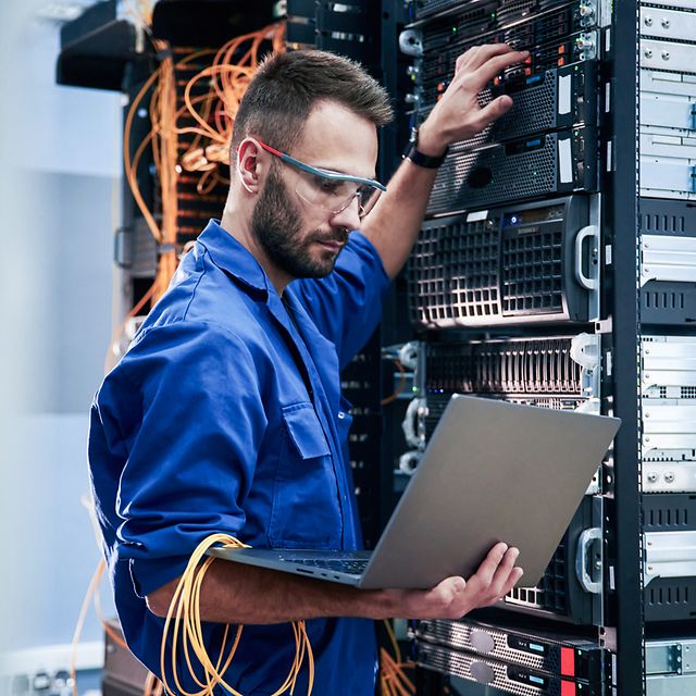 Junger Mann mit Laptop arbeitet mit Internetgeräten und Kabeln im Serverraum