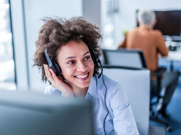 Frau mit wuscheligen Haaren sitzt lächelnd mit Headset in einem Büro