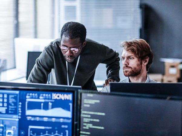 Zwei männliche Kollegen arbeiten im vier-Augen-Prinzip vor zwei Monitoren