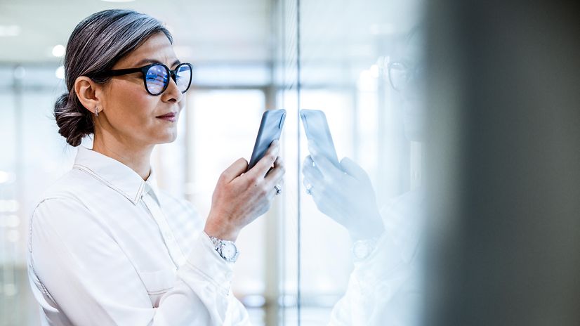 Lächelnde Frau steht vor einem Bürofenster mit einem einem Smartphone in der Hand