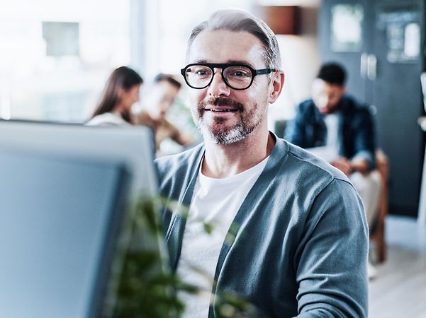 Mann im Büro am Computer, im Hintergrund ein Teammeeting
