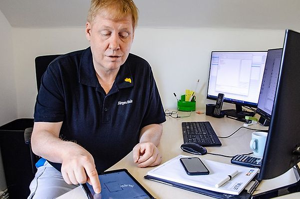Malermeister Juergen Hahn im blauen T-Shirt mit tippt auf Tablet an seinem Büroschreibtisch