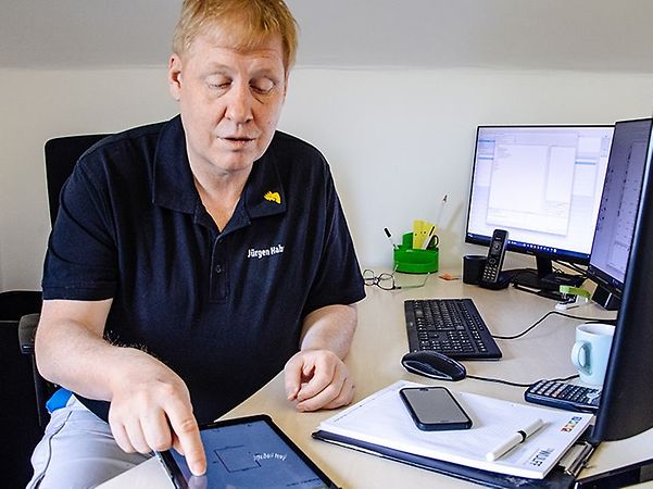 Malermeister Juergen Hahn im blauen T-Shirt mit tippt auf Tablet an seinem Büroschreibtisch