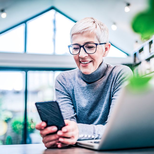 Eine lächelnde Frau die auf ihr Smartphone schaut, während sie am Schreibtisch mit Laptop sitzt.