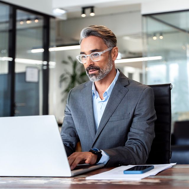 Mann mit grauem Haar und Brille arbeitet im Büro am Laptop, Dokumente und Smartphone auf dem Schreibtisch.