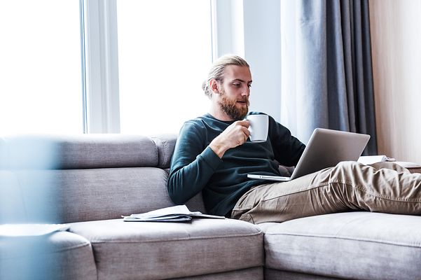 Konzentrierter junger bärtiger Mann sitzt zu Hause und benutzt einen Laptop.