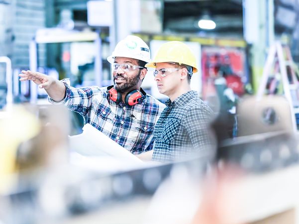 Zwei Arbeiter mit Schutzhelmen und Schutzbrillen unterhalten sich in einer Fabrikhalle
