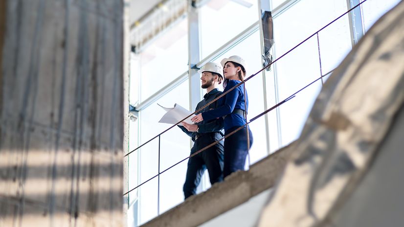 Frau und Mann in Businesskleidung mit Bauhelmen stehen in einer Fabrikhalle