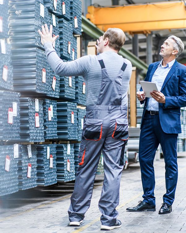 Business Analytics Zwei Männer stehen in einer Lagerhalle vor mehreren Paletten von Rohren