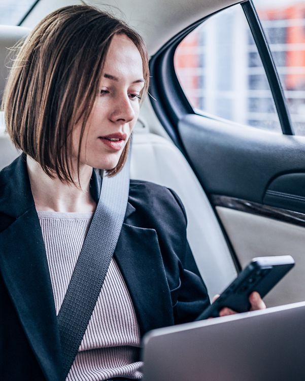 Business Analytics Frau sitzt in Ihrem Auto hinten und arbeitet an Ihrem Laptop und schaut auf Ihr Smartphone