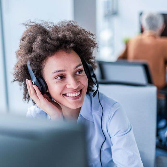 Frau mit wuscheligen Haaren sitzt lächelnd mit Headset in einem Büro