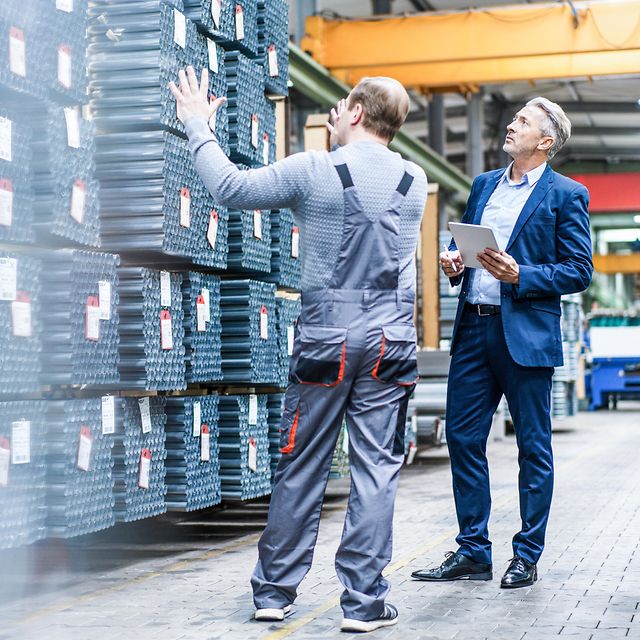 Business Analytics Zwei Männer stehen in einer Lagerhalle vor mehreren Paletten von Rohren