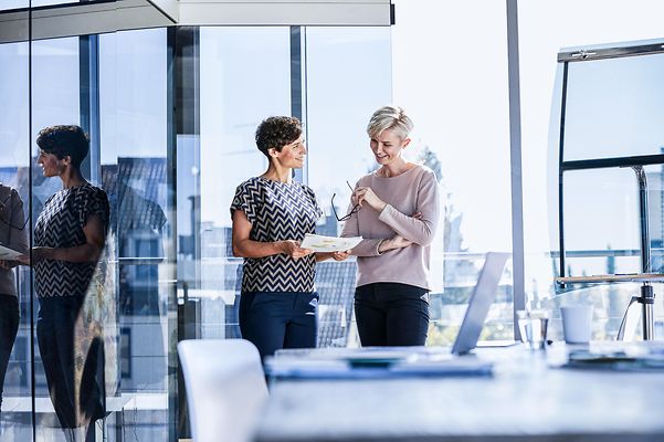 Zwei lächelnde Geschäftsfrauen diskutieren Dokumente am Fenster im Büro