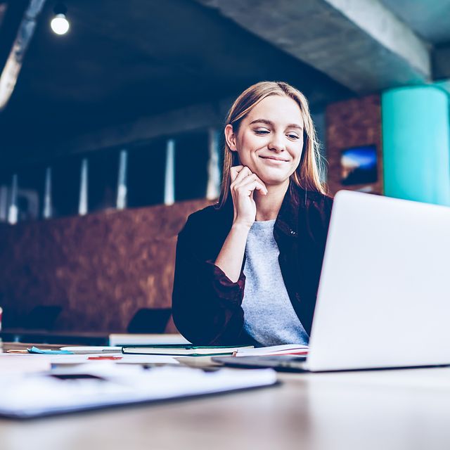 Eine Frau sitzt in einem modernen Büro an einem Tisch mit Unterlagen und schaut auf den Bildschirm.
