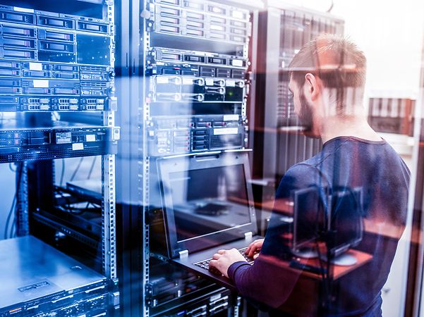 Ein junger Mann, der einen Laptop hält, steht im Serverraum und arbeitet an einem Server