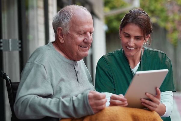 Pflegerin und älterer Mann im Rollstuhl schauen gemeinsam auf ein Tablet